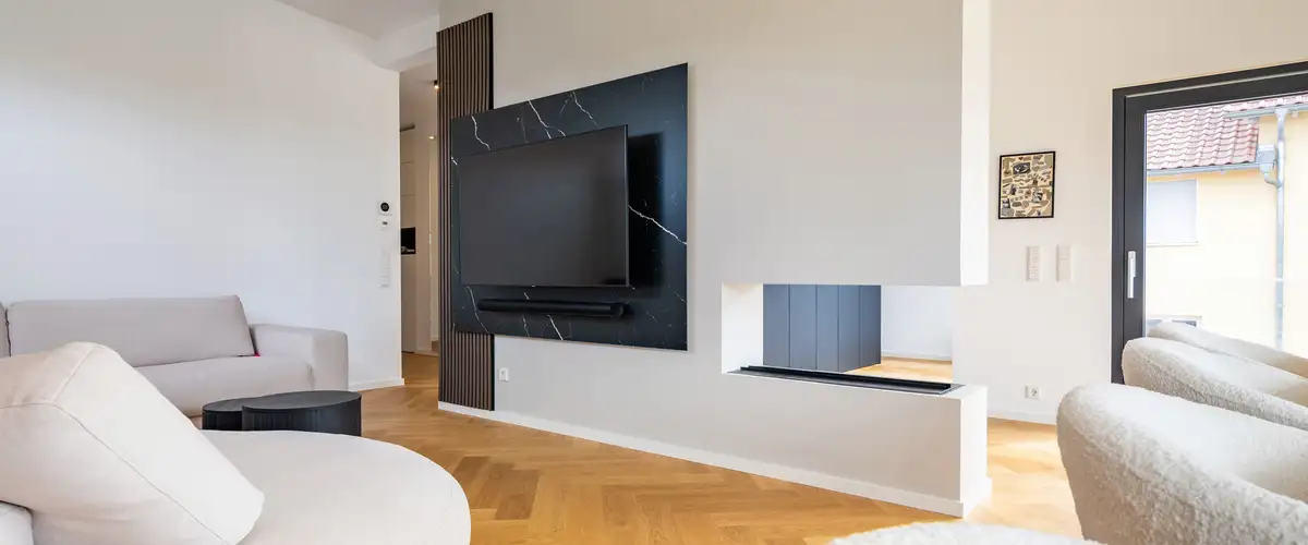 Wohnbereich mit Fischgrätenparkett, beiger Couch, Blick auf den Flachbildfernseher am Raumtrennerelement