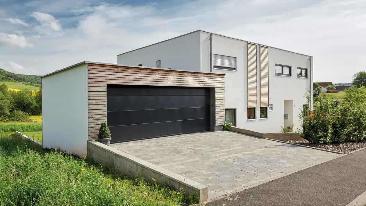 Flachdachhaus in leichter Hanglage mit der Zufahrt von oben, im Bild links mit Garage, holzverkleidete Garage mit grauem Garagentor